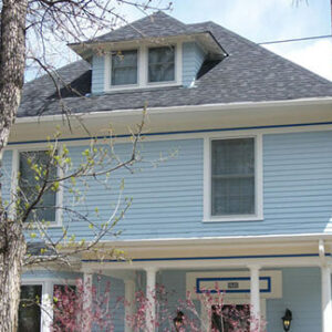 A light blue two story house with white trim, a gray shingled roof, a dormer window, a front porch, and a tree with pink blossoms in front.