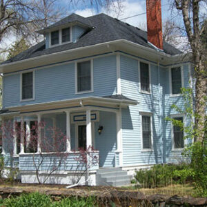 A two story light blue house with white trim, a covered front porch, gabled roof, brick chimney, and surrounding trees under a blue sky.