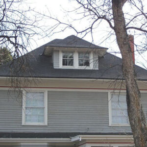 Upper portion of a two story house with gray siding, a dark gray hipped roof, a dormer window, a brick chimney, and bare tree branches in front