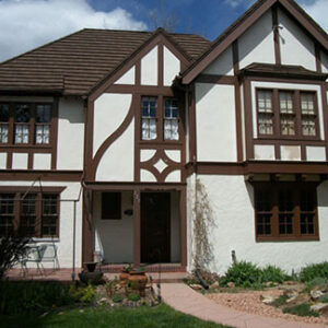 A Tudor style house with white stucco walls, dark brown wood trim, a tiled roof, multi pane windows, a front door, and a landscaped yard with a stone path and rock garden under a blue sky.