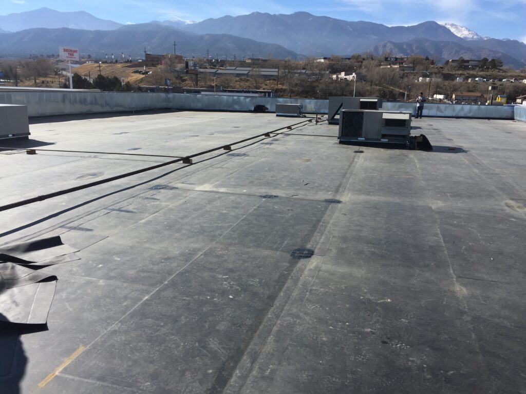 A flat commercial rooftop with rubber membrane roofing, HVAC units, a worker standing near the edge, and a mountain range and cityscape visible in the background.