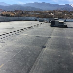 A flat commercial roof with membrane roofing, HVAC units, and pipes, overlooking a town with mountains in the background.