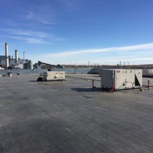 A flat commercial rooftop with membrane surface, HVAC and rooftop mechanical units, conduit piping, and an industrial facility with smokestacks visible in the background under a blue sky.