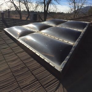 A metal skylight or solar panel structure with domed glass sections mounted on a shingled roof, with bare trees and hills visible in the background.