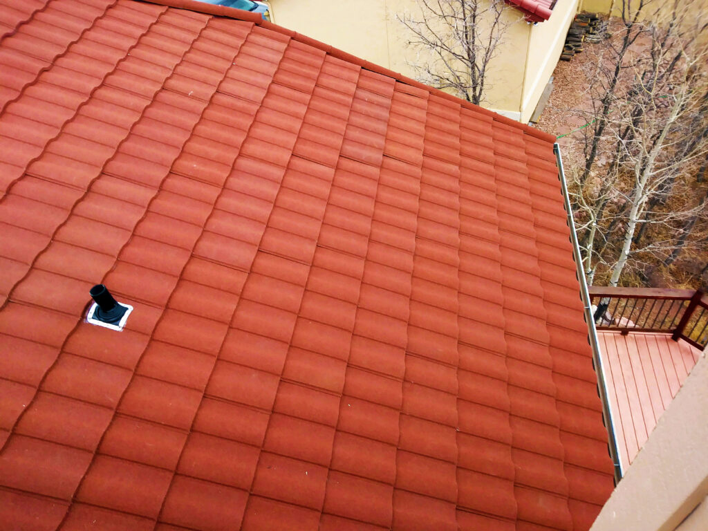 Aerial view of a red tile roof with a plumbing vent pipe, overlooking a wooden deck and bare trees below.