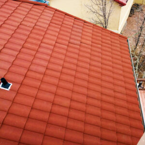 A red tiled roof viewed from above with a small vent pipe and a yellow building wall and bare trees in the background.