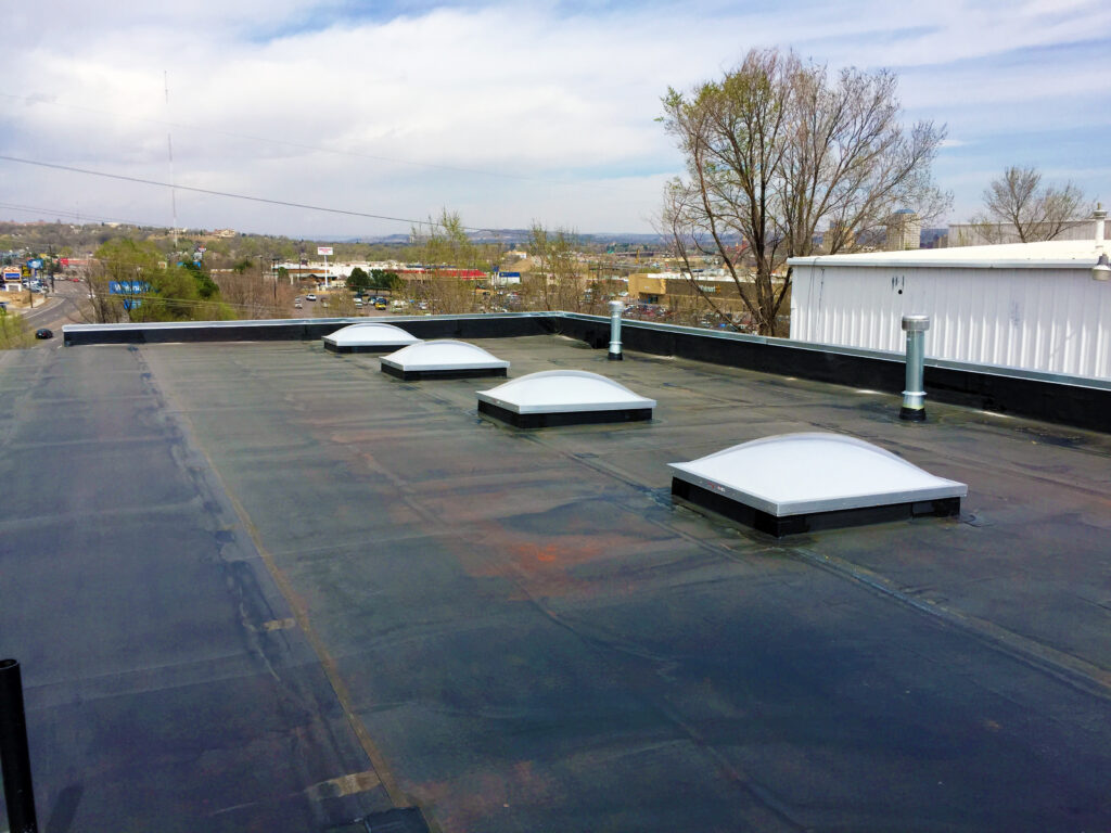 A flat commercial roof with four domed skylights and roof vents, overlooking a parking lot, trees, and a distant cityscape under a partly cloudy sky.