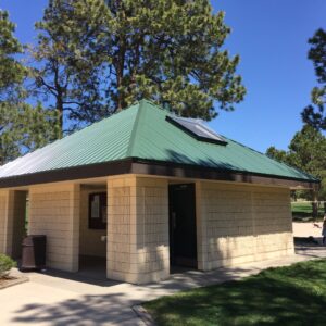A small park restroom building with a green metal roof and tan block walls, surrounded by tall pine trees, grass, and a paved walkway under a clear blue sky.