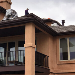 A worker installing shingles on a house roof, with a stucco exterior, balcony railing, and windows visible below under an overcast sky.