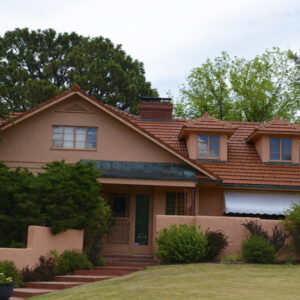 A two story stucco house with a terracotta tile roof, dormer windows, a chimney, an arched front door with an awning, and a landscaped front yard with shrubs, trees, and a brick walkway.