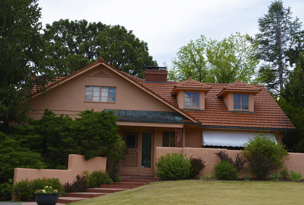 A stucco house with a red tile roof, dormer windows, brick chimney, front porch entrance, low garden wall, and surrounding trees and shrubs, with a green lawn in front.