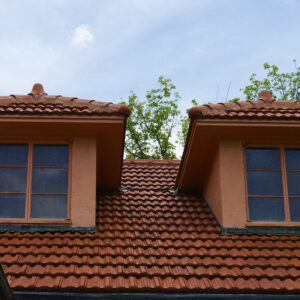 A terracotta tile roof with two dormer windows and stucco walls, trees and sky visible in the background.