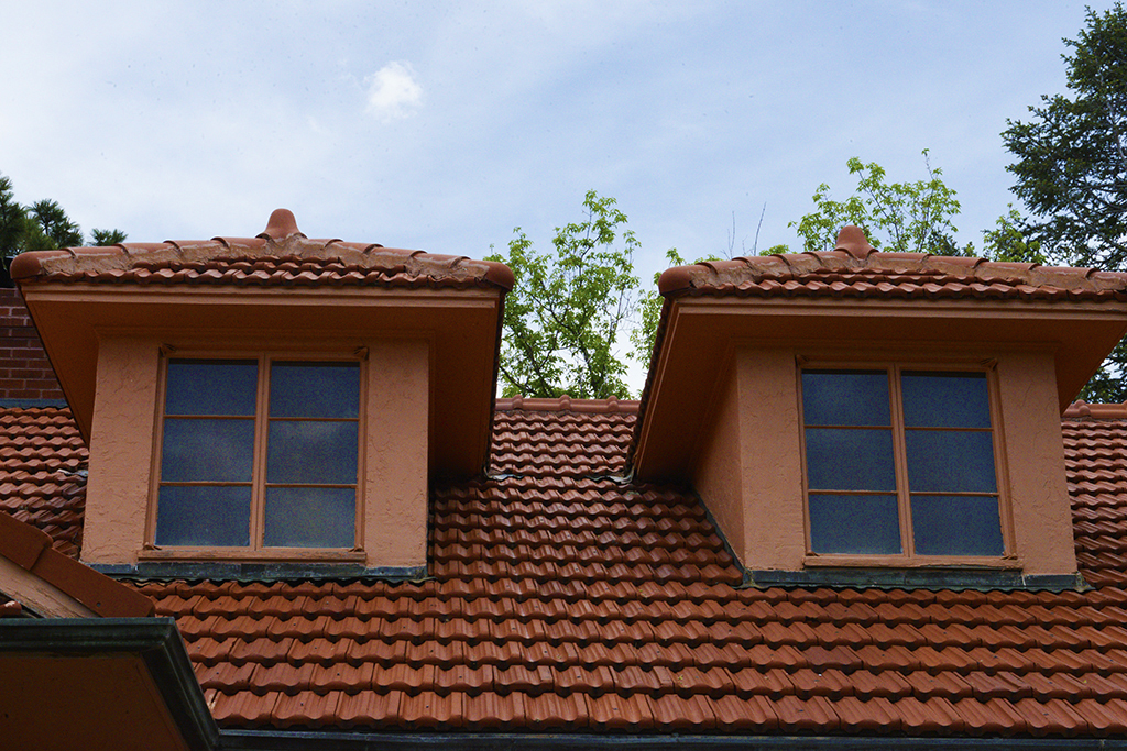 A red tiled roof with two stucco dormer windows, framed by trees and a partly cloudy sky.