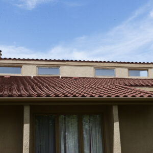 Exterior view of a stucco building with a red tiled roof, small upper windows, and a curtained window below, set against a blue sky.