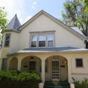 A two story stucco house with a turret, arched porch entrance, gabled roof, and surrounding trees under a blue sky.