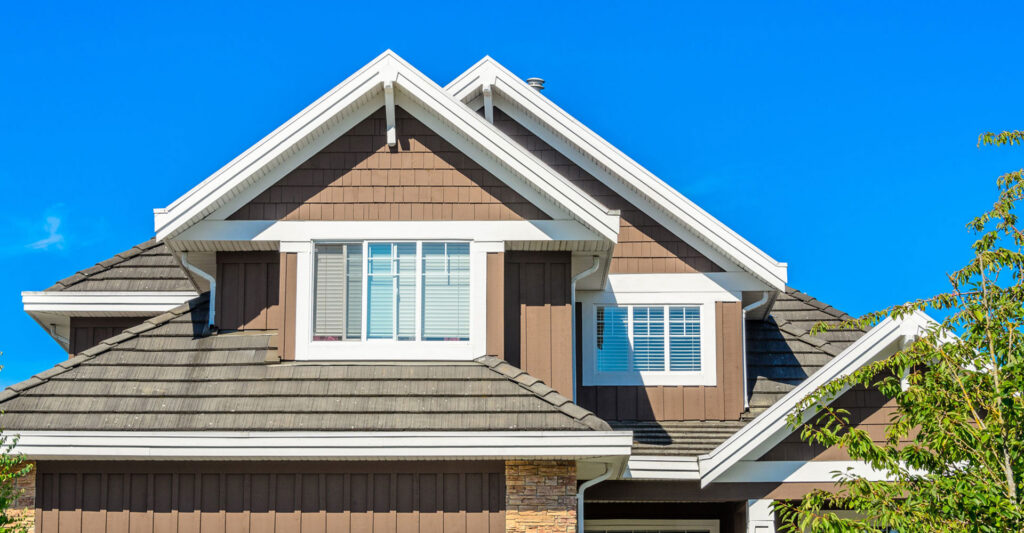 Upper facade and gabled roofline of a house with brown shingle siding, white trim, dormer windows with blinds, and a tiled roof against a clear blue sky with a tree branch at the edge.